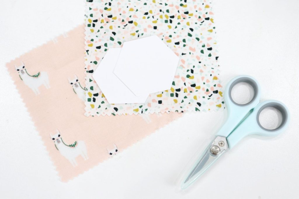 Image contains two 5” squares of fabric, two paper hexagon shapes, and a pair of scissors on a white background.