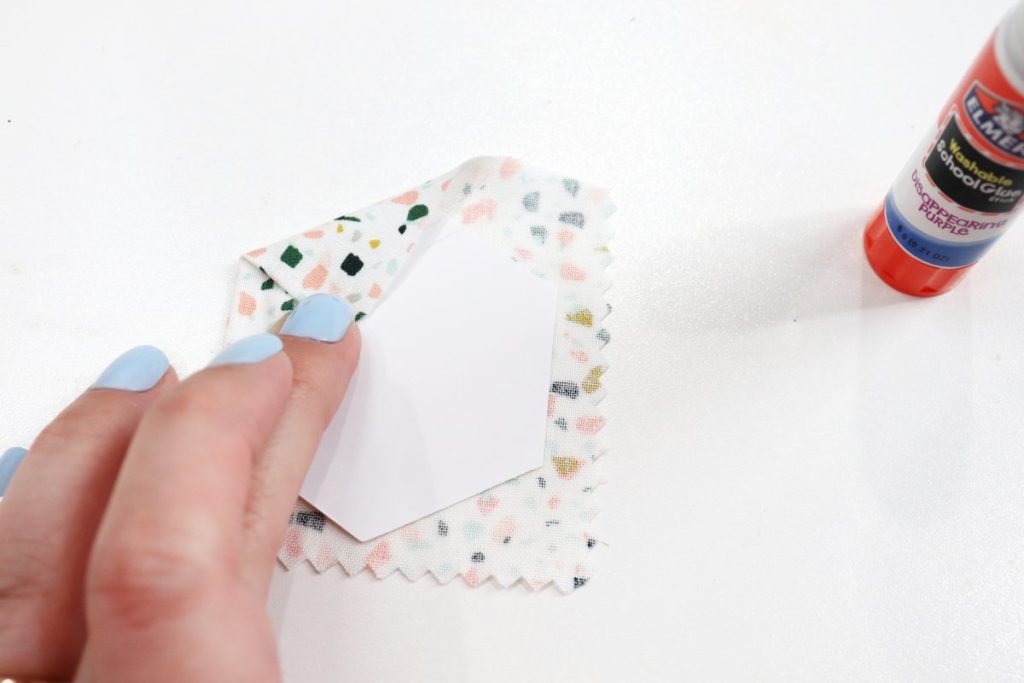 Image contains Amy’s hand wrapping one side of a fabric square around a paper hexagon. A glue stick is off to the side on a white background.