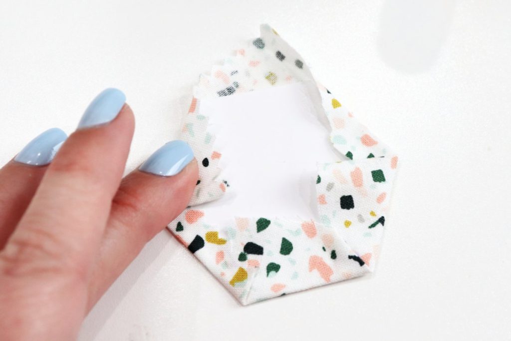 Image contains Amy’s hand wrapping five sides of a paper hexagon with fabric on a white background.