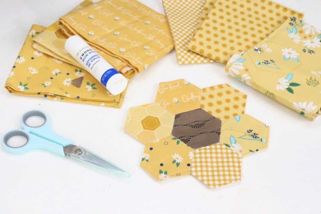 Image contains the fabric sunflower coaster and a pair of scissors in the foreground, and assorted yellow fabrics and glue stick in the background on a white table.