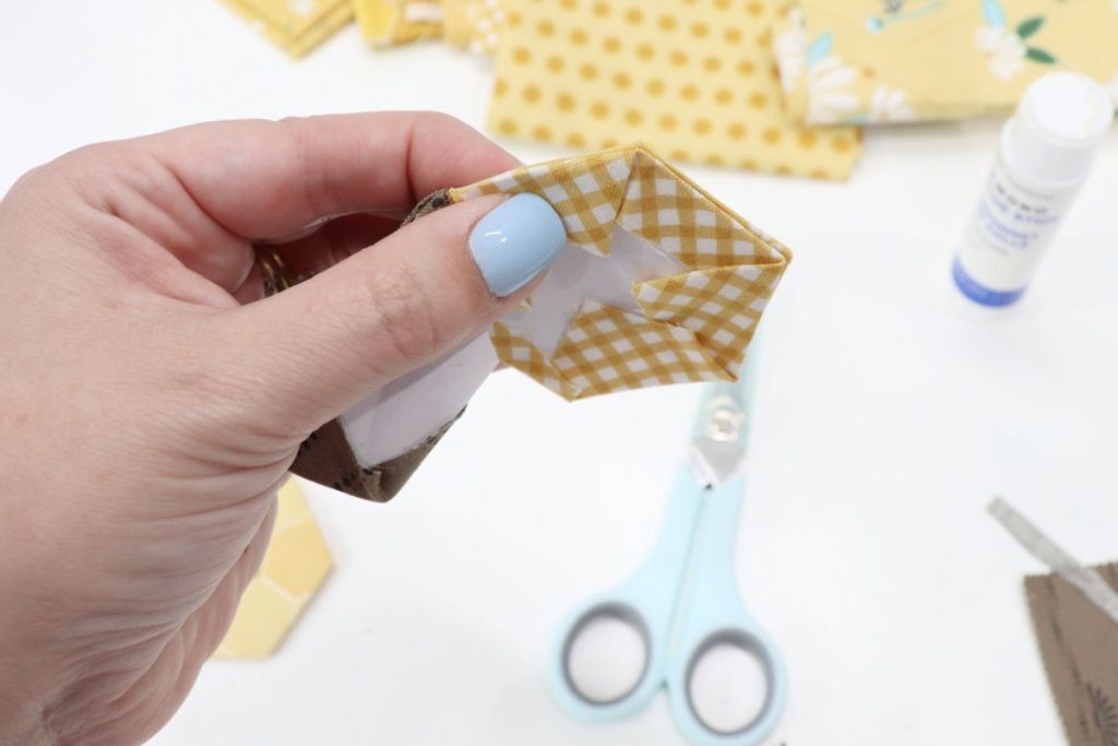 Image contains Amy’s hand holding three fabric hexagons that are being sewn together. In the background, yellow fabric, a glue stick, and scissors sit on a white table.