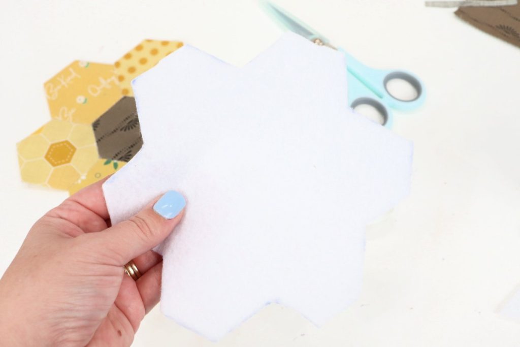 Image contains Amy’s hand holding a piece of white felt cut to match the shape of the sunflower. The fabric flower and a pair of scissors sit on a white table in the background.