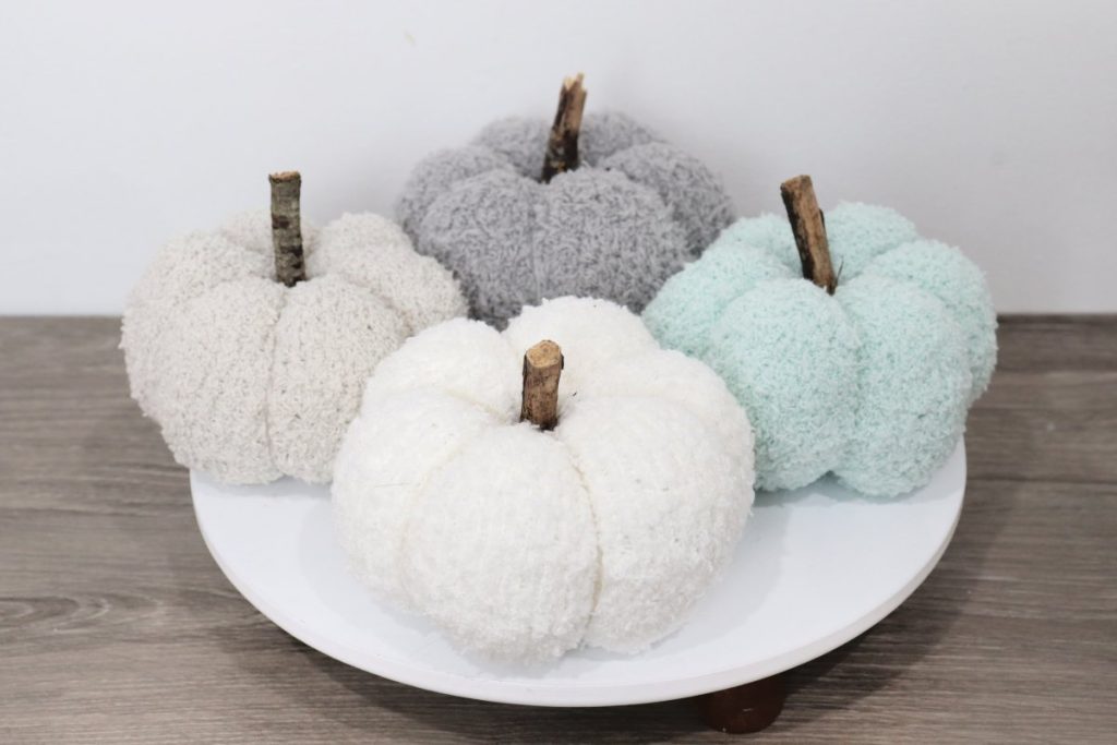 Image contains four pumpkins made from fuzzy socks; a tan, a grey, a mint, and a cream one, sitting on a wooden desk.