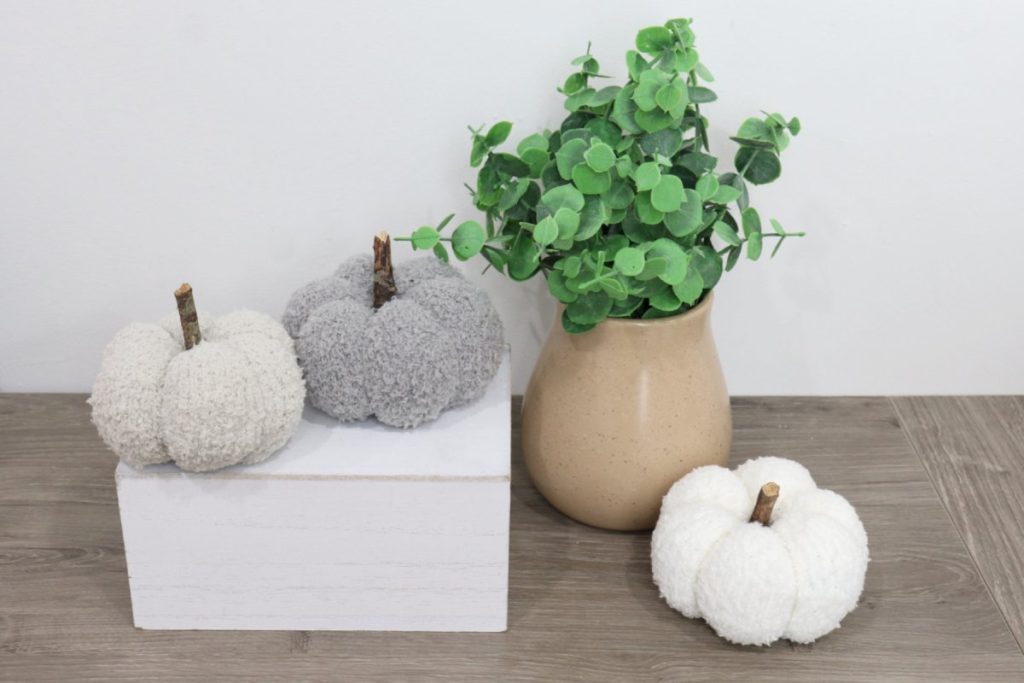 Image contains three sock pumpkins: one tan, one grey, and one cream, next to a faux plant on a wooden desk.