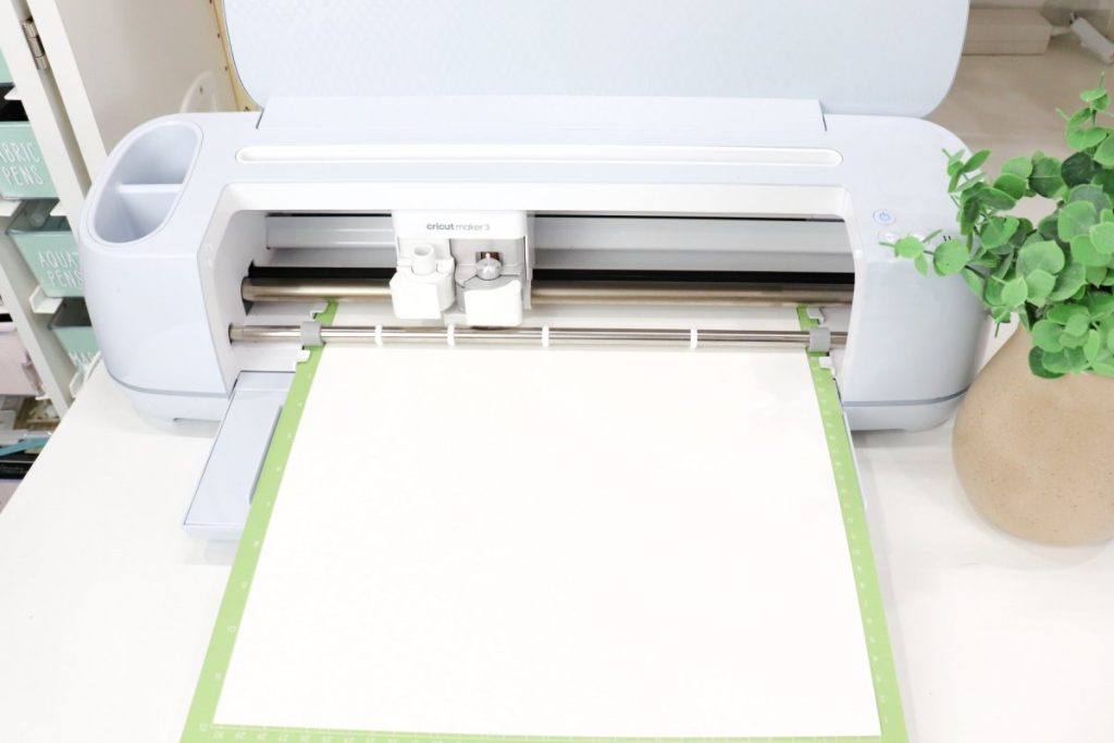 Image contains a pale blue Cricut Maker 3 machine with a piece of white cardstock loaded into it on a green cutting mat.