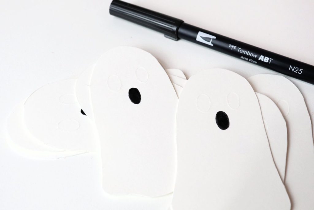 Image contains a pile of white ghost shapes cut from cardstock, with black ovals colored in for the mouths. A black marker sits nearby.