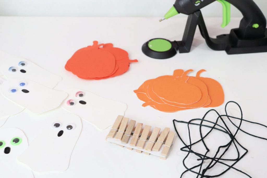 Image contains a white workspace covered with orange cardstockc pumpkins, white cardstock ghosts with google eyes, small clothespins, black twine, and a glue gun.