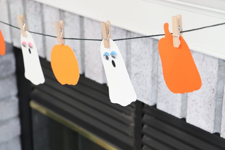 Image contains a close-up view of the finished Boo Garland, featuring white cardstock ghosts and orange cardstock pumpkins attached to black twine with small wooden clothespins.