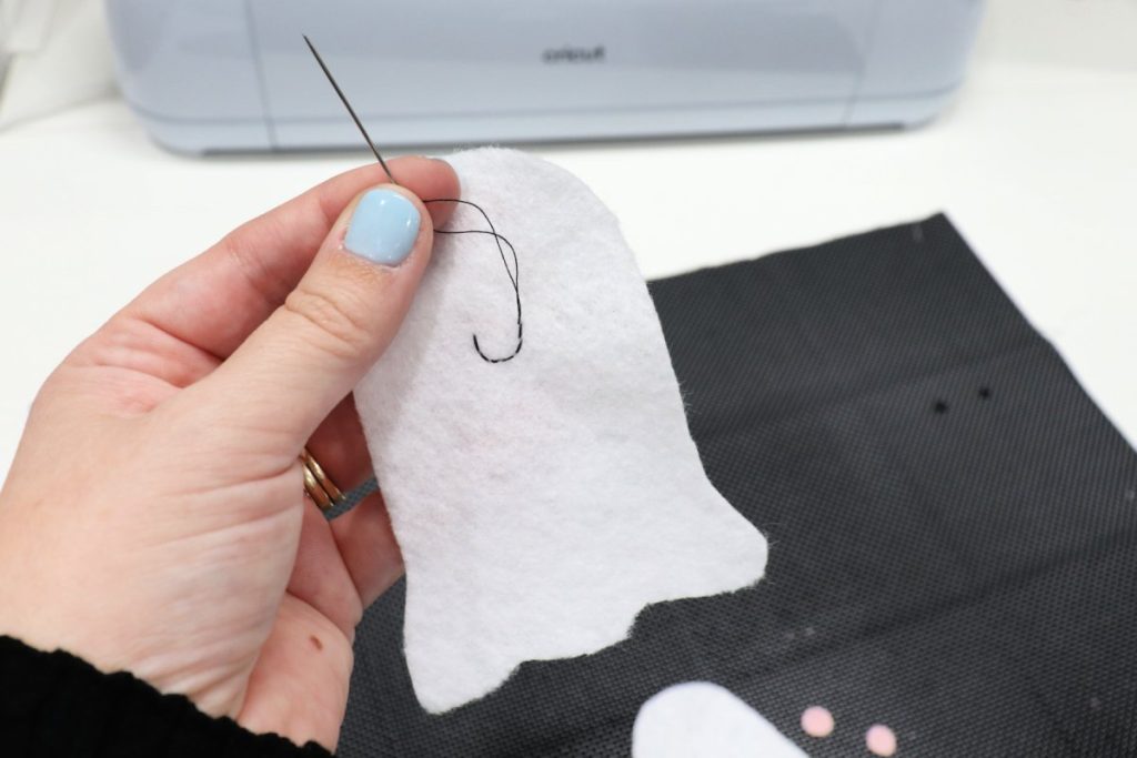 Image contains Amy’s hand holding a white felt ghost shape and a sewing needle. A black smile is stitched onto the felt shape.