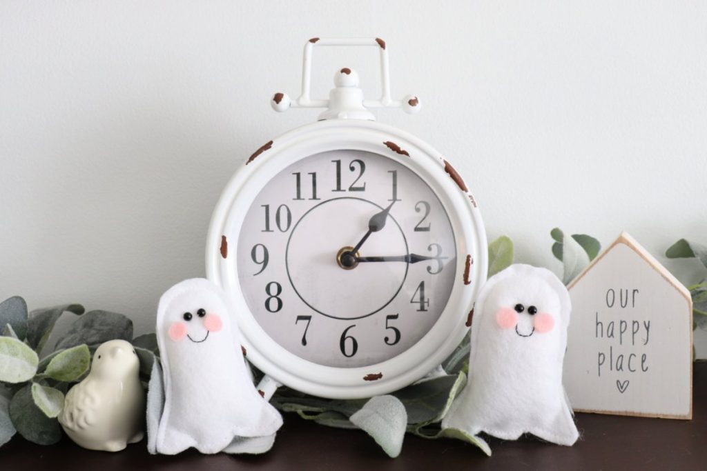 Image contains a white mantel clock with a small felt ghost sitting on each side of it. Other decor, like a eucalyptus garland, a porcelain bird, and a wooden house shape are in the background.