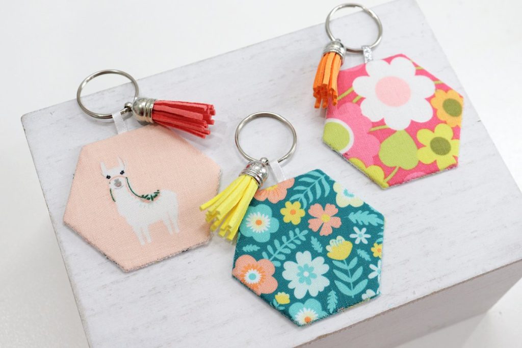 Image contains three fabric keychains; one blush with a llama, one teal with multicolored flowers, and one hot pink with multicolored flowers.