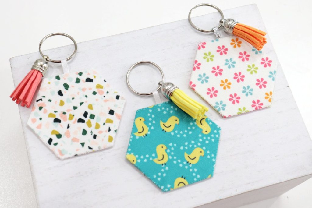 Image contains the backs of three fabric keychains with tassels. One has a multicolored terracotta pattern, one is teal with yellow ducks, and one is white with small multicolored flowers.
