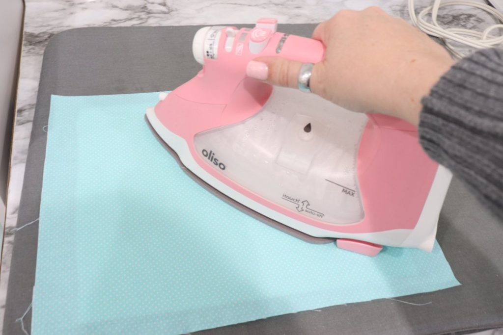 Image contains Amy’s hand holding a pink Oliso iron and ironing a piece of teal backing fabric.