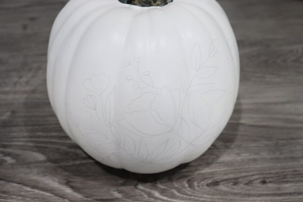 Image contains a white pumpkin with a floral design drawn on in pencil, sitting on a wooden desk.
