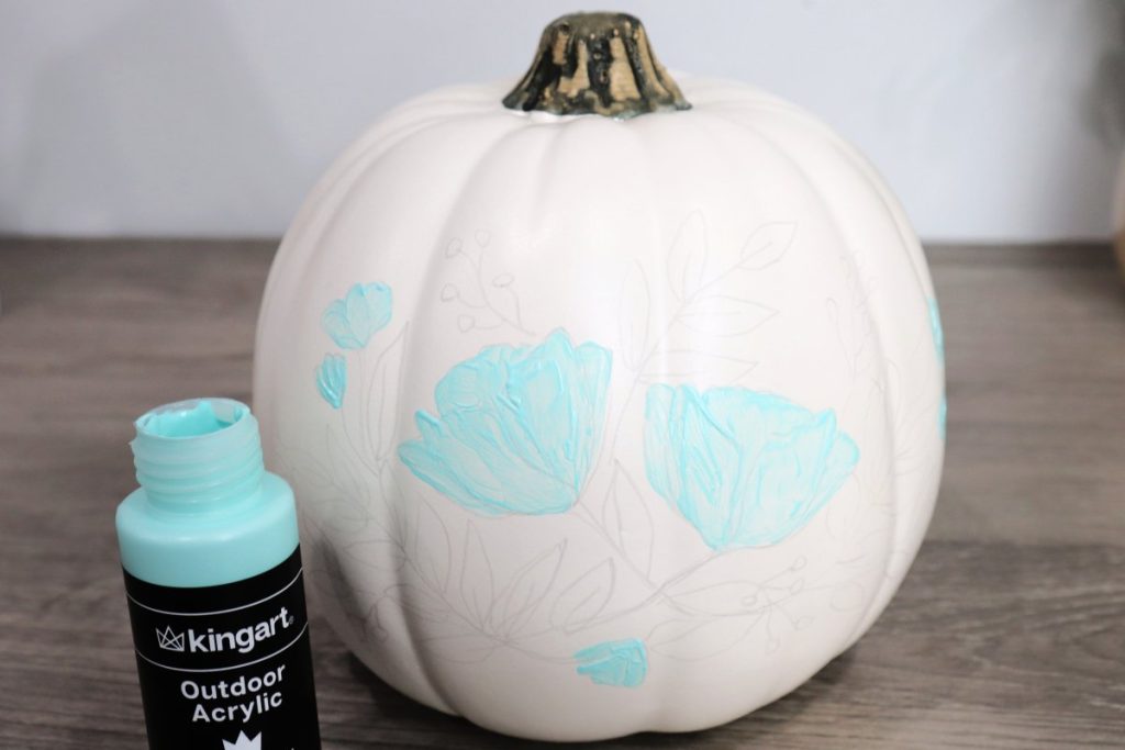 Image contains a white pumpkin with a penciled floral design on a wooden desk. The large flowers are painted teal.
