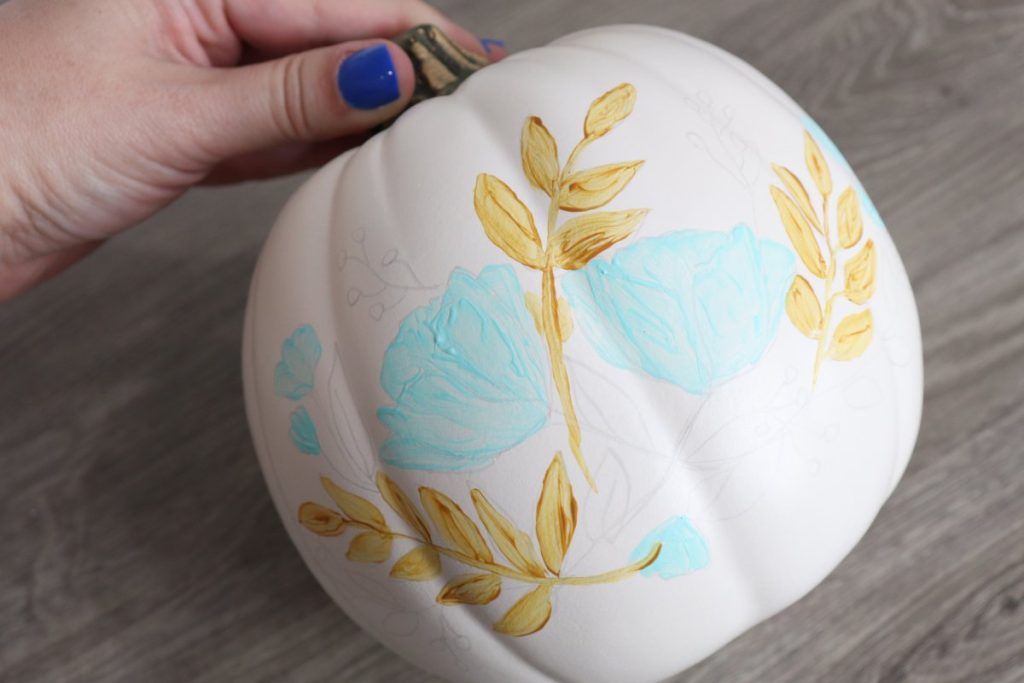 Image contains Amy’s hand holding a white pumpkin by the stem. There are teal flowers and yellow leaves painted, and the rest of the design is drawn in pencil.