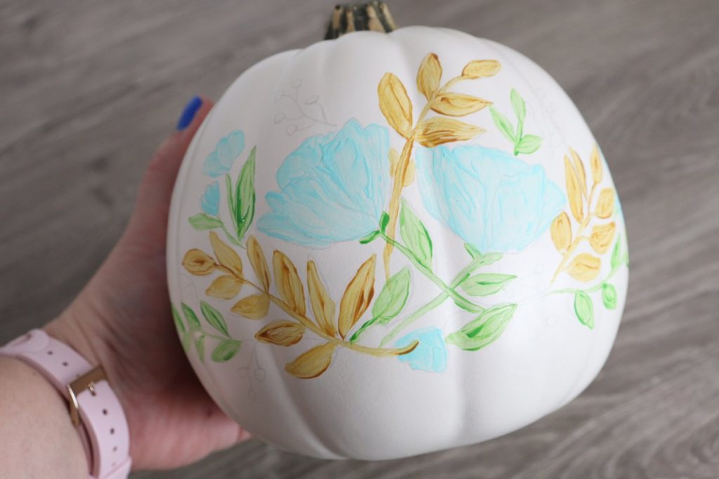 Image contains Amy’s hand holding a white pumpkin with teal flowers and green and yellow leaves painted on the front. The rest of the design is sketched lightly in pencil.