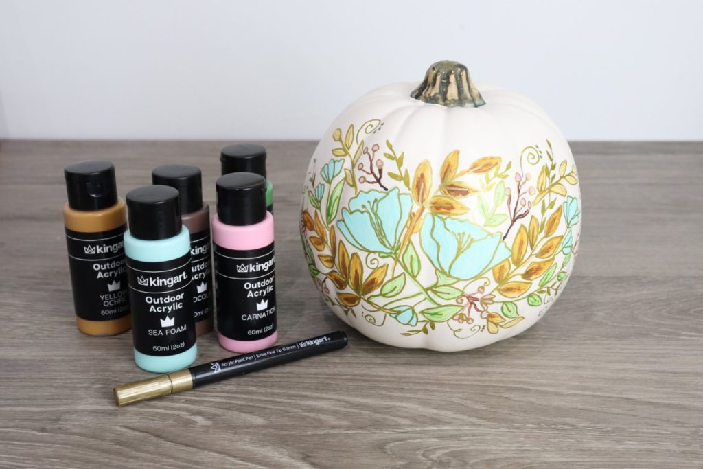 Image contains a white pumpkin with a floral design sitting on a wooden desk next to five bottles of acrylic paint and a gold paint marker.