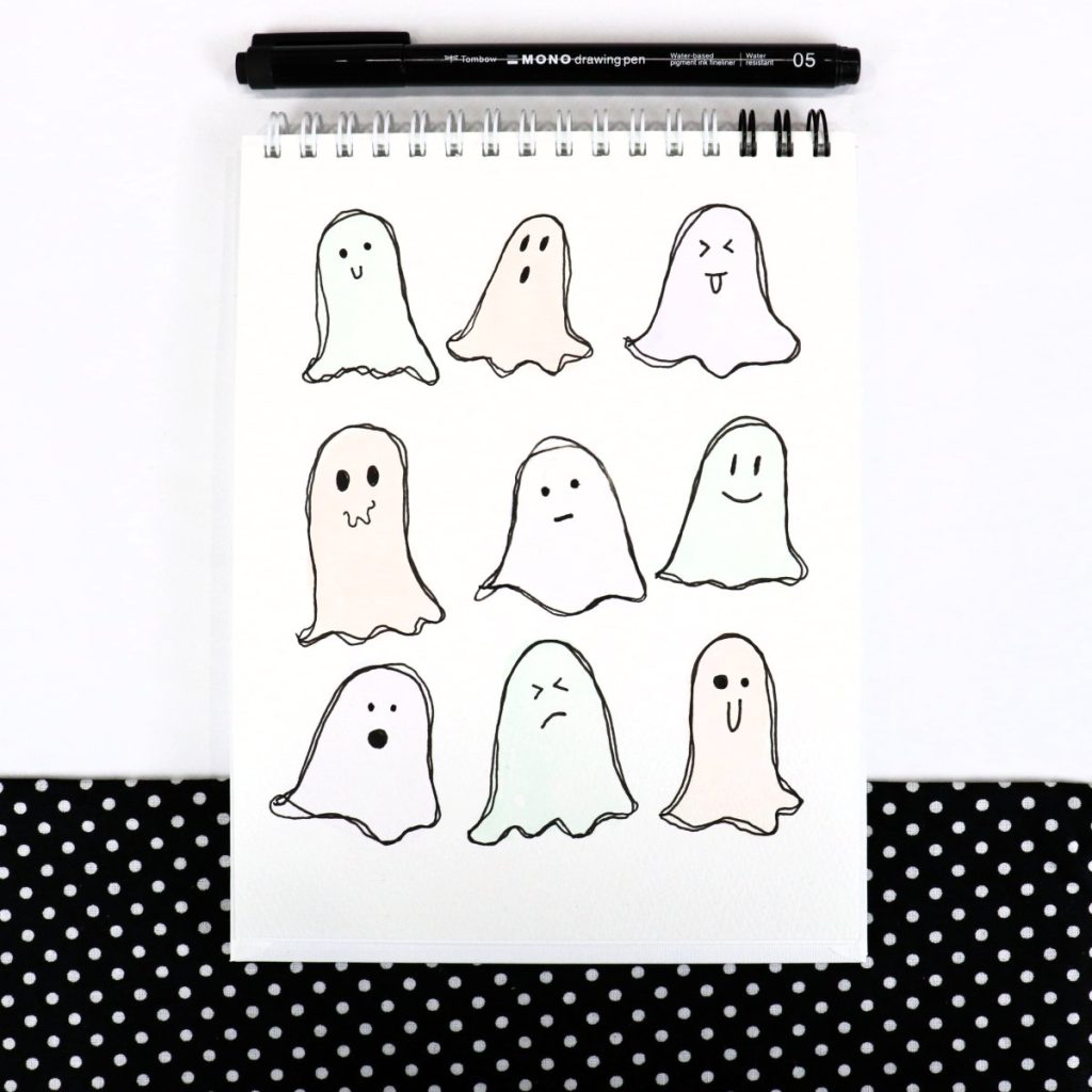 Image contains an open sketch book with nine pastel colored ghosts drawn on the page. A black drawing pen sits above it on a white table with a piece of black and white polka dot fabric.