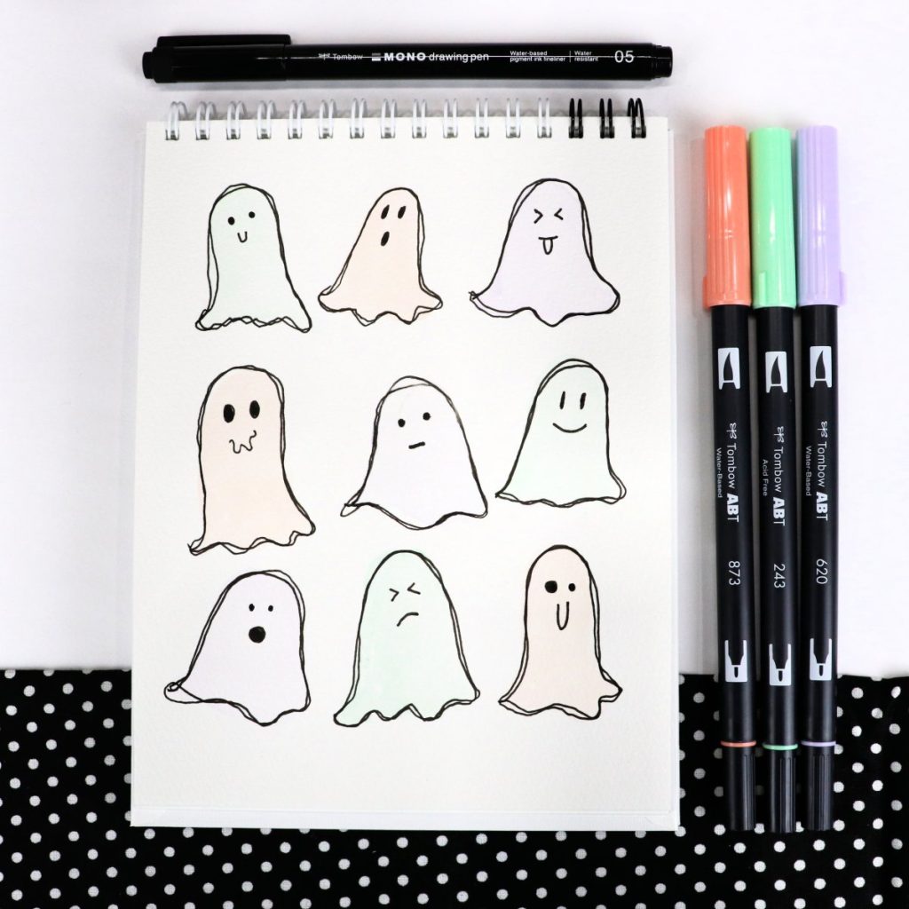 Image contains an open sketchbook with nine pastel colored ghosts drawn on the top page. Each has a different facial expression. A MONO Drawing Pen sits above the sketchbook, and three Dual Brush Pens (orange, green, and purple) sit off to the right.