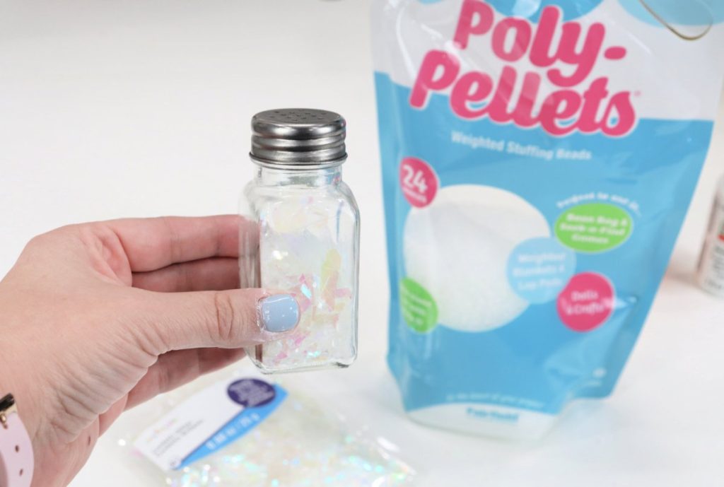 Image contains Amy’s hand holding a clear glass salt shaker filled with glitter and Poly Pellets. The bags of glitter and pellets are in the background on a white table.