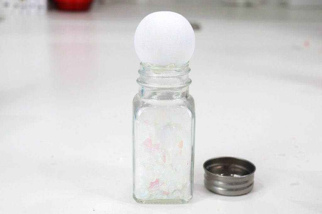 Image contains a clear glass salt shaker filled with glitter and Poly Pellets, with a painted white wooden bead on top. The cap is removed and sits to the side.