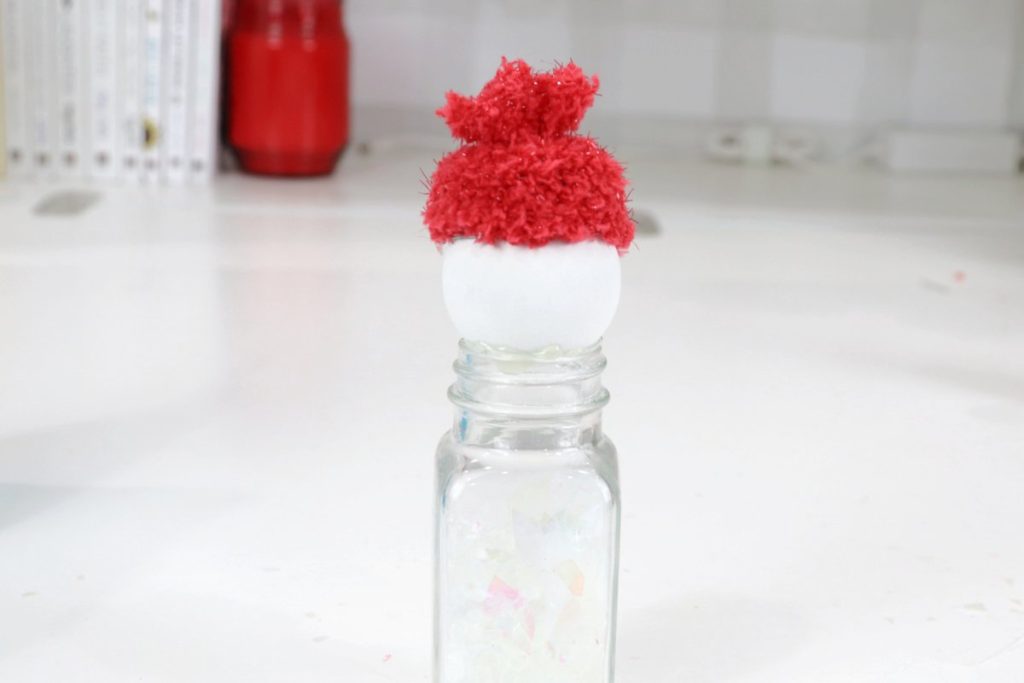 Image contains a clear salt shaker filled with glitter and Poly Pellets, that has a white wooden bead and a fuzzy red hat on top.