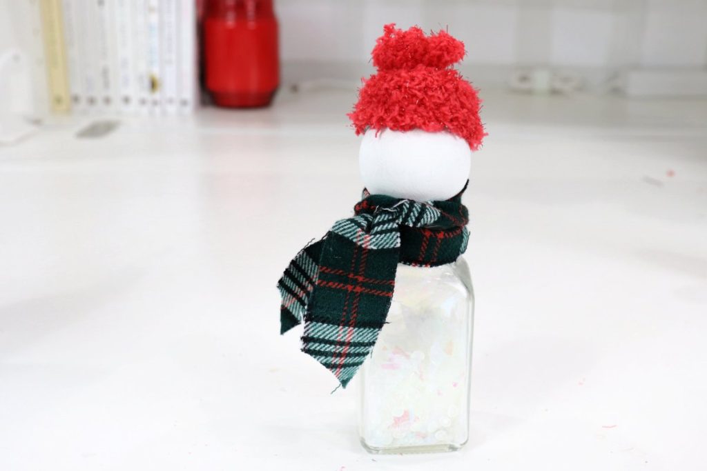 Image contains a salt shaker filled with glitter that has a white wooden bead for a head and a red hat on top. The area below the head is wrapped with a plaid flannel scarf.