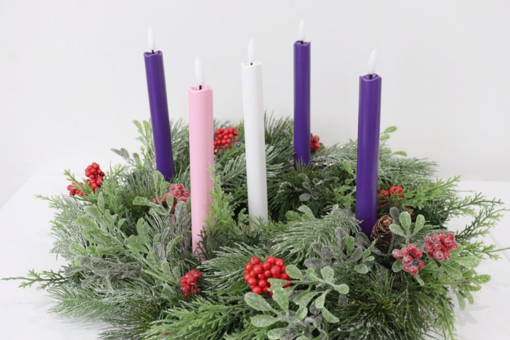 Image contains a wreath made from five types of faux greenery. It also has clusters of red berries; some with a glittered snow effect. Three purple candles and one pink candle are inserted into the wreath, and a white candle stands in the center.