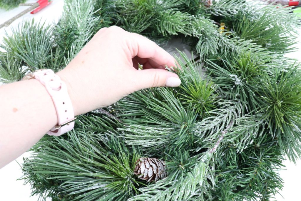 Image contains Amy’s hand inserting a short piece of faux pine greenery into a wreath made from three types of greens.
