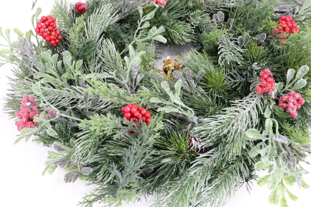 Image contains a wreath made from five types of faux greenery. It also has clusters of red berries; some with a glittered snow effect.