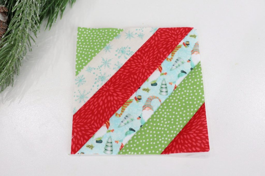 Image contains the top of the striped fabric coaster, trimmed to be a 5” square. It is made of assorted red, green, white, and teal holiday fabrics.