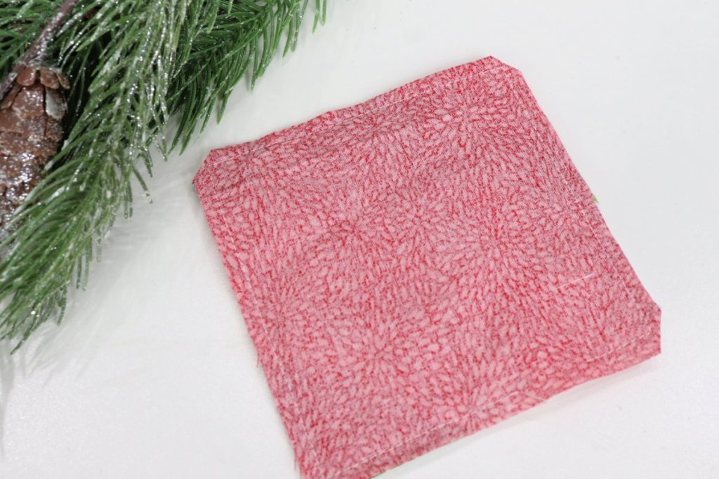 Image contains a 5” red square of fabric laying face down on top of the coaster top. A pine branch sits nearby.