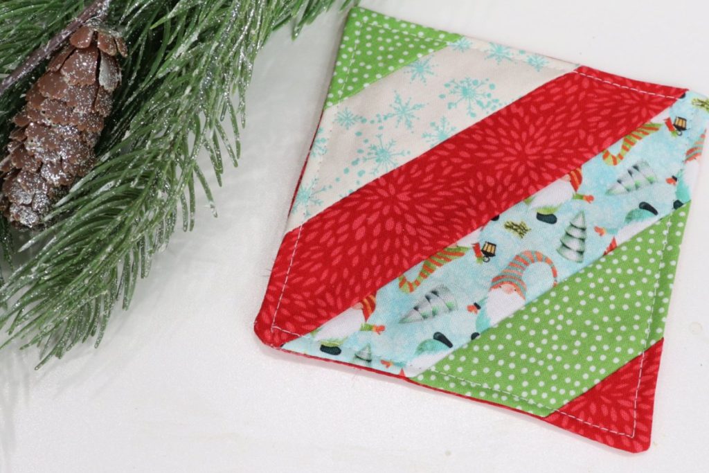 Image contains a diagonally striped coaster made from assorted holiday fabrics. A pine branch sits next to it on a white background.