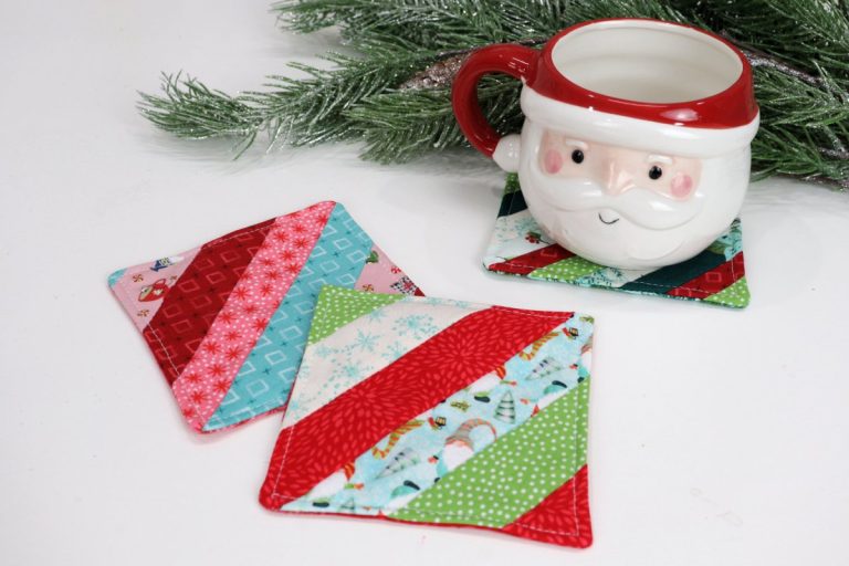 Image contains two striped fabric coasters made from assorted holiday fabric scraps. Behind them, a santa mug sits on a third fabric coaster. A faux pine branch is in the background.