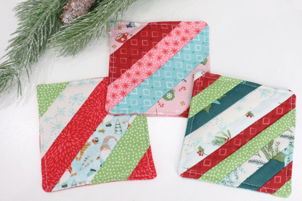 Image contains three striped fabric coasters made from a variety of red, green, white, teal, and pink holiday fabrics. A pine branch sits nearby.