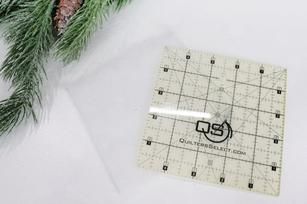 Image contains a 5” square quilting template ruler on top of a 5” square of white batting. A faux pine branch sits nearby.