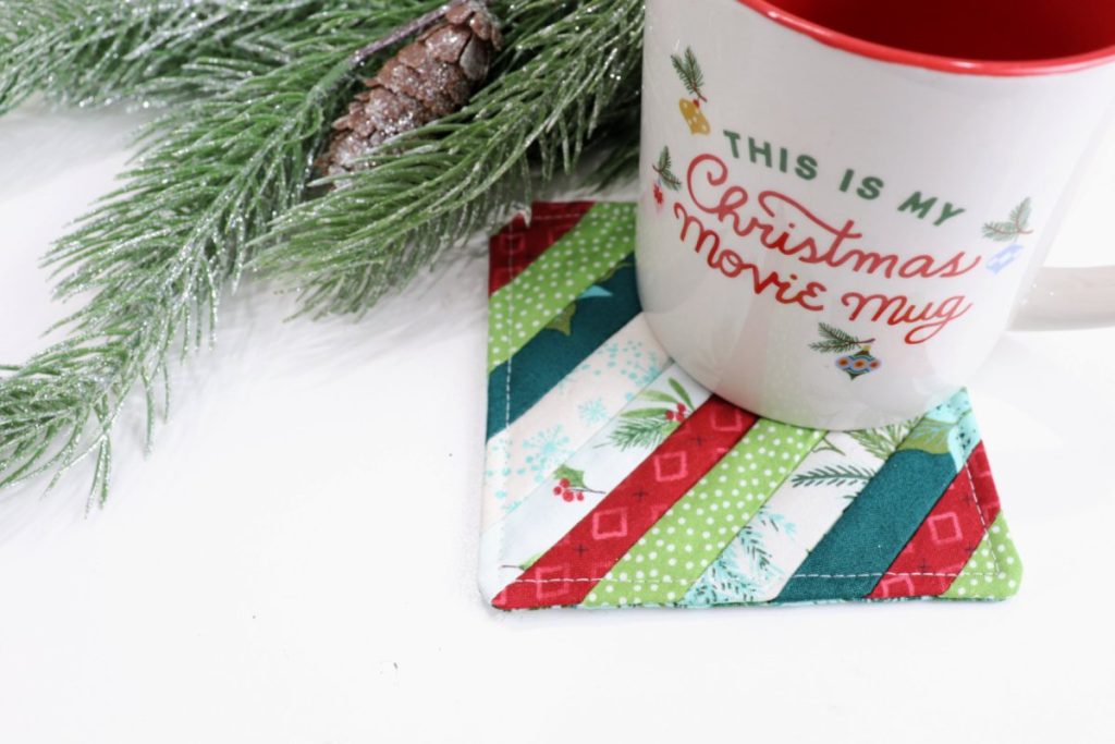 Image contains a white mug that reads, “this is my Christmas movie mug,” sitting on a striped fabric coaster made from assorted red, green, and white holiday fabrics. A faux pine branch sits in the background.