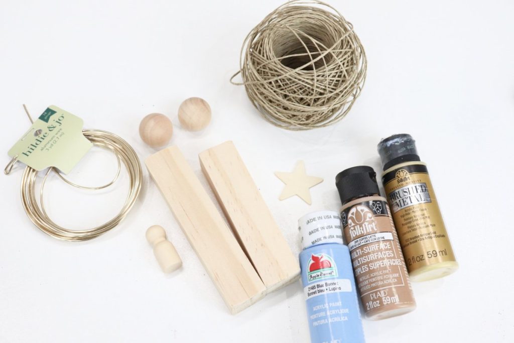 Image contains a roll of gold wire, two wooden blocks, two wooden beads, a wooden star, and a wooden peg person, a roll of twine, and three bottles of acrylic paint (blue, brown, gold) on a white background.