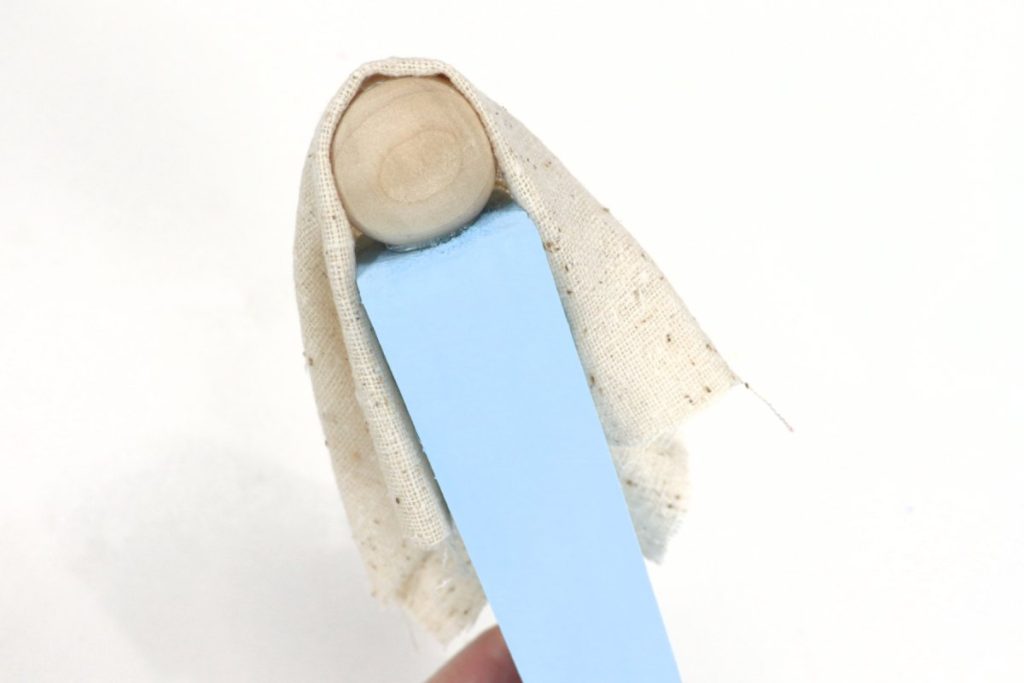 Image contains a blue wooden block with a natural wooden bead head and a neutral linen head covering draped over the bead.