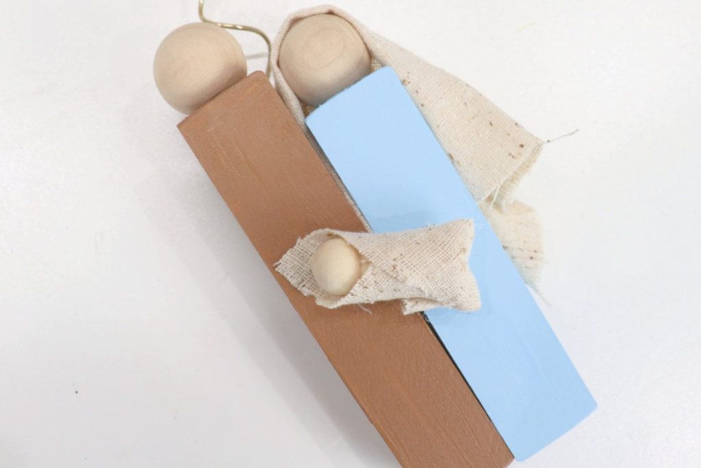 Image contains brown and blue blocks with wooden bead heads that have been glued together along one side. The fabric-wrapped peg person is glued on top of the blocks.