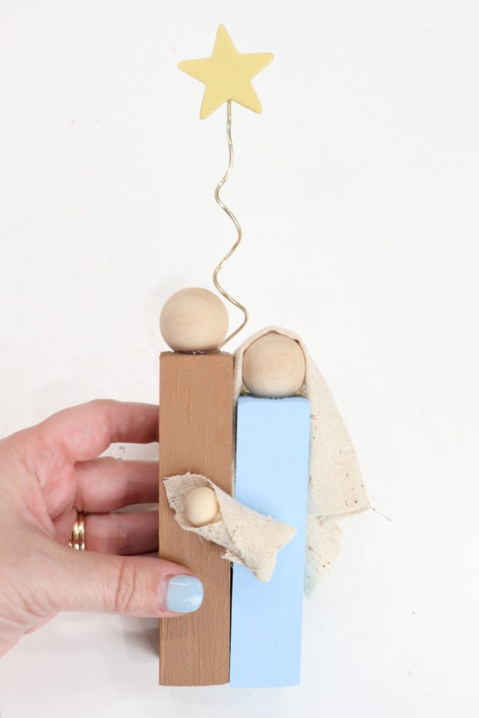 Image contains Amy’s hand holding holy family made of wooden blocks and beads on a white background.