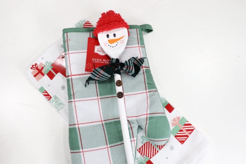 Image contains a wooden spoon snowman on top of a green plaid oven mitt and a white dish towel with printed red, pink, and green packages.