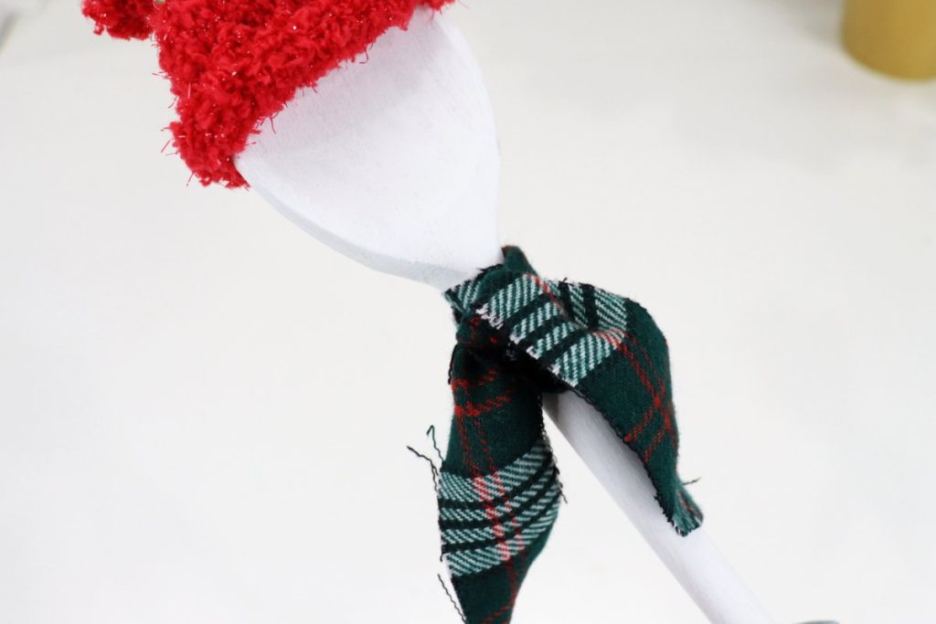 Image contains a white wooden spoon with a red fabric hat, and a green and red plaid flannel scarf tied around the handle.