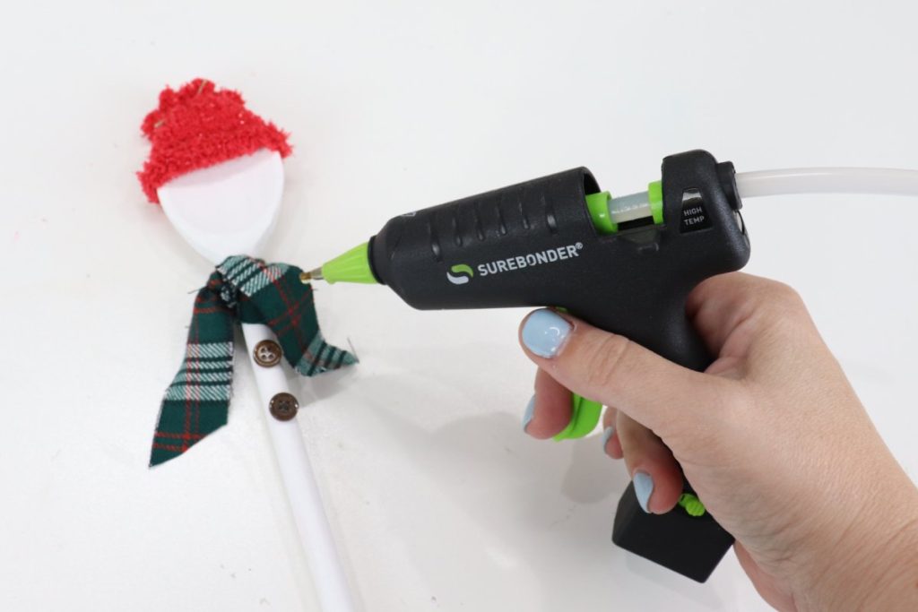 Image contains Amy’s hand holding a black Surebonder mini glue gun and adding two brown buttons to the wooden spoon snowman’s handle.