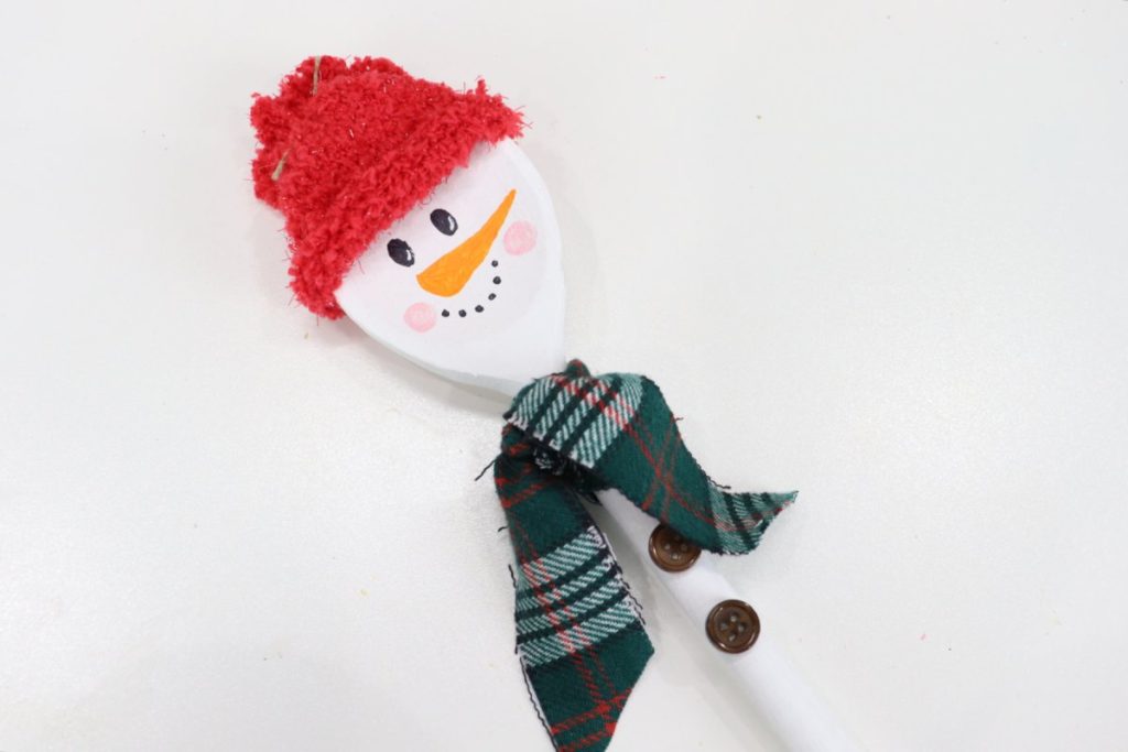 Image contains a wooden spoon snowman with a red fuzzy hat, a plaid flannel scarf, two brown buttons, and a cheerful face.