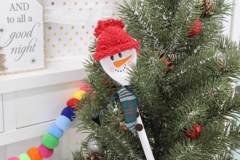 Image contains a snowman made from a wooden spoon, with a red fuzzy hat and a plaid flannel scarf. It is in front of an artificial Christmas tree.