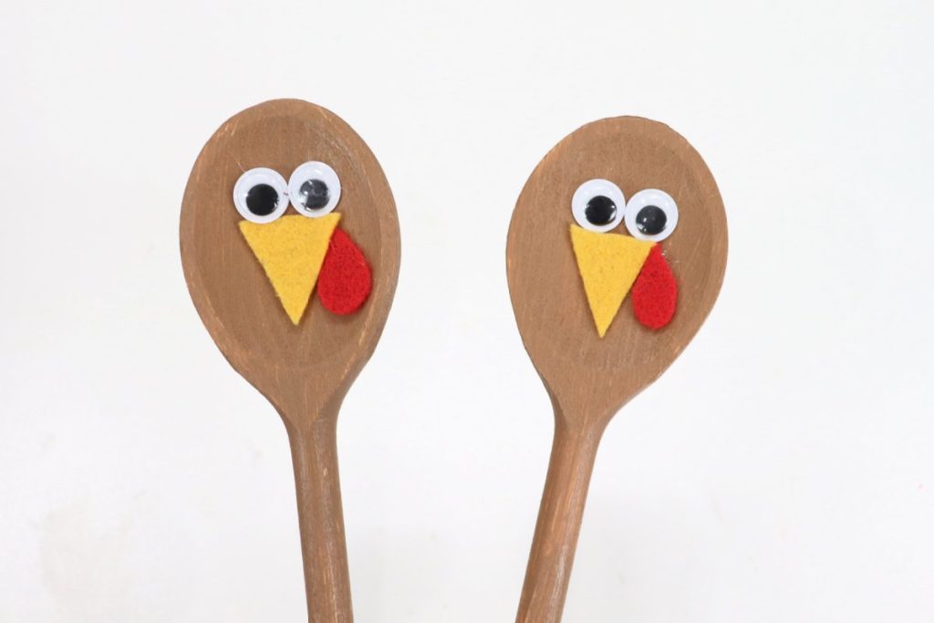 Image contains the tops of two wooden spoons that have been painted brown. Each one has a pair of googly eyes, a yellow felt beak, and a red felt snood.