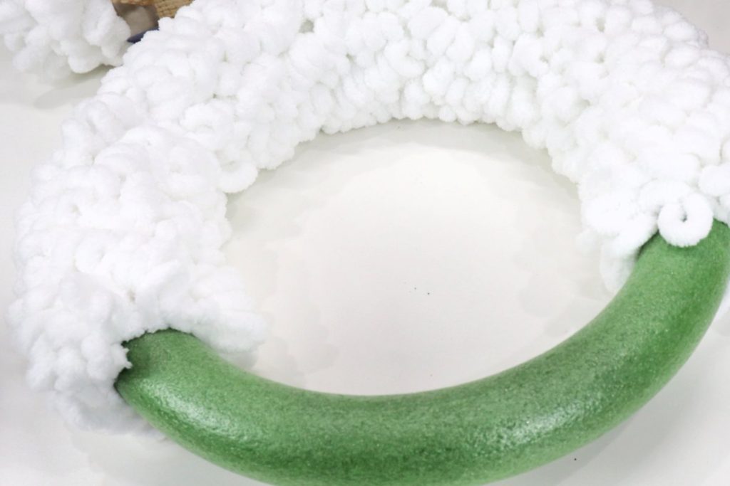 Image contains a green styrofoam wreath form half wrapped with loopy white yarn on a white table.