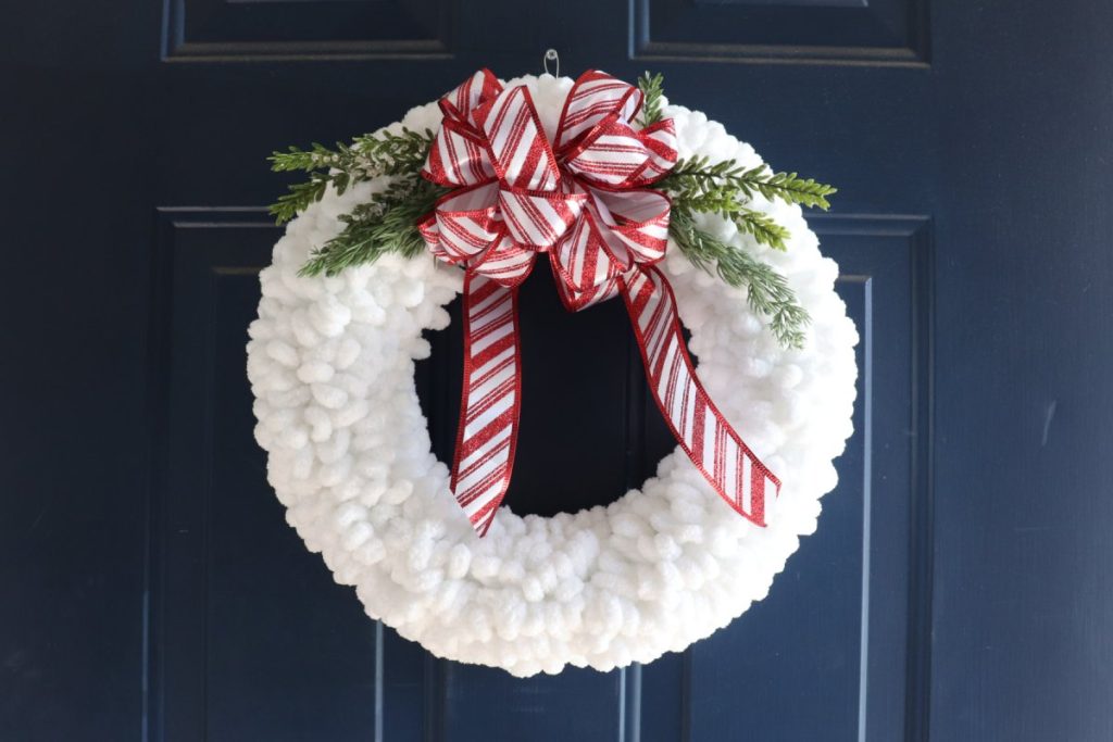 Image contains a wreath made of loopy white yarn with a red and white bow and faux greenery on a dark blue door.